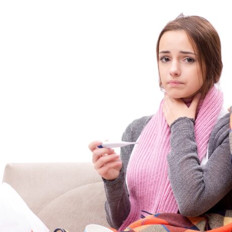 O adolescentă care stă pe canapea cu durere în gât și termometru în mână, sugestiv pentru mononucleoza infecțioasă care afectează adolescenții și adulții tineri